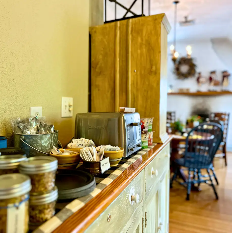 Breakfast buffet setup with jars, utensils, and a toaster on a counter, leading to a dining area with wooden chairs and a fireplace in the background.