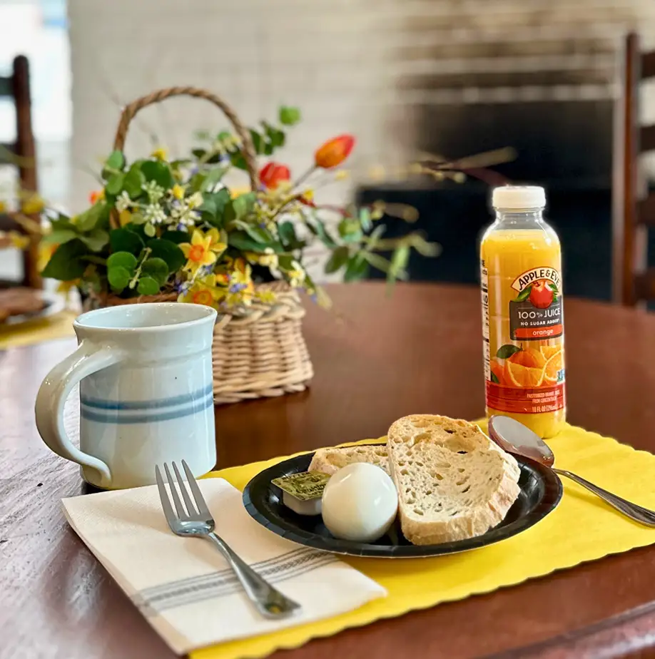 Breakfast table with a plate of bread and a boiled egg, a bottle of juice, and a coffee cup, set in a cozy room with a wreath and colonial figurines on the fireplace mantel.