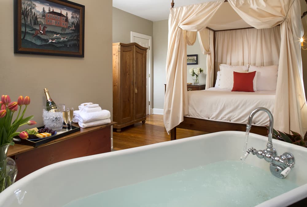 A spacious bedroom with hardwood floors, a canopy bed, an inviting bathtub, and a tray of cheese, fruit and champagne on a sideboard.