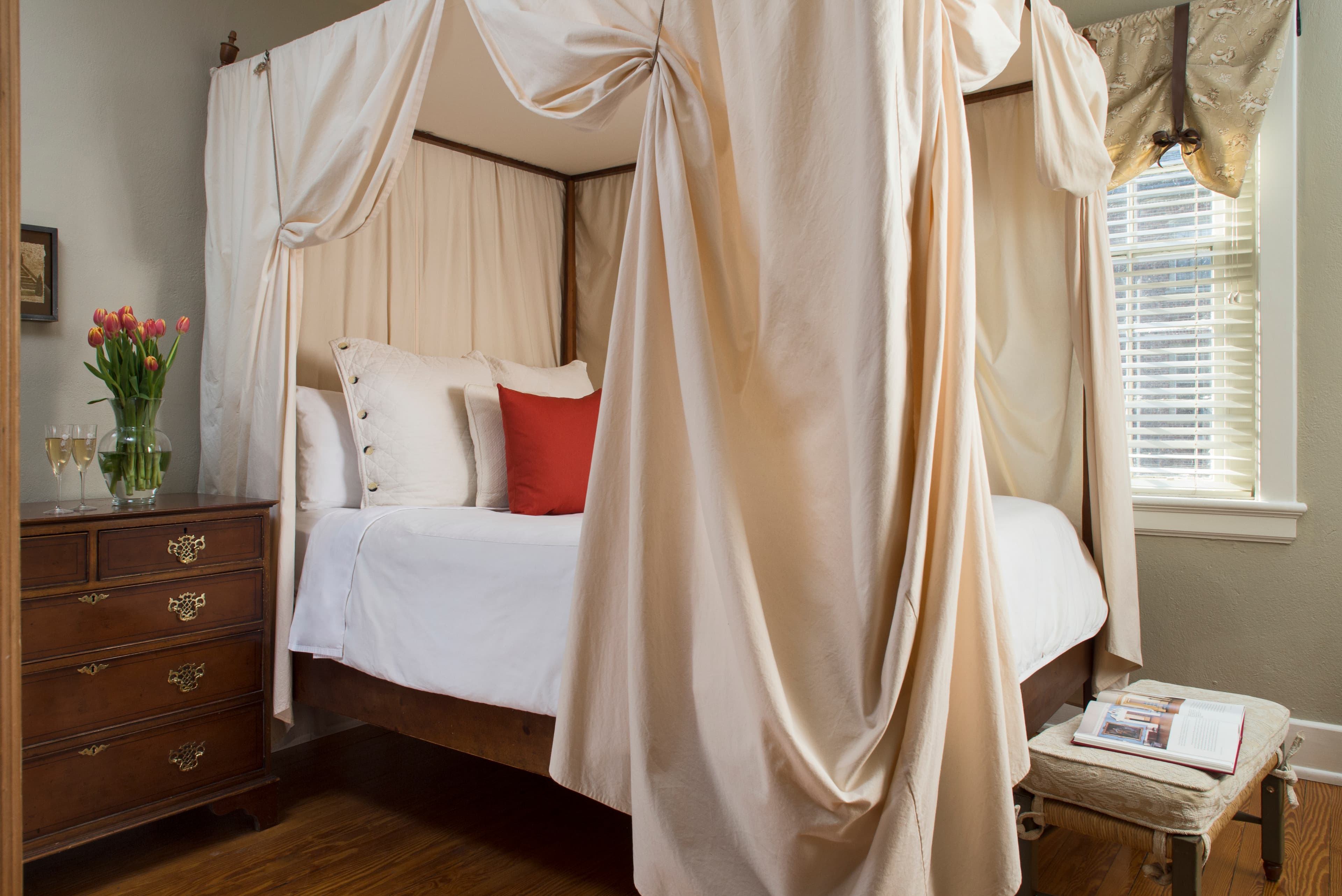 Heavily draped four-poster canopy bed with decorative pillows, bureau with vase of flowers, bench with cushion at foot of bed