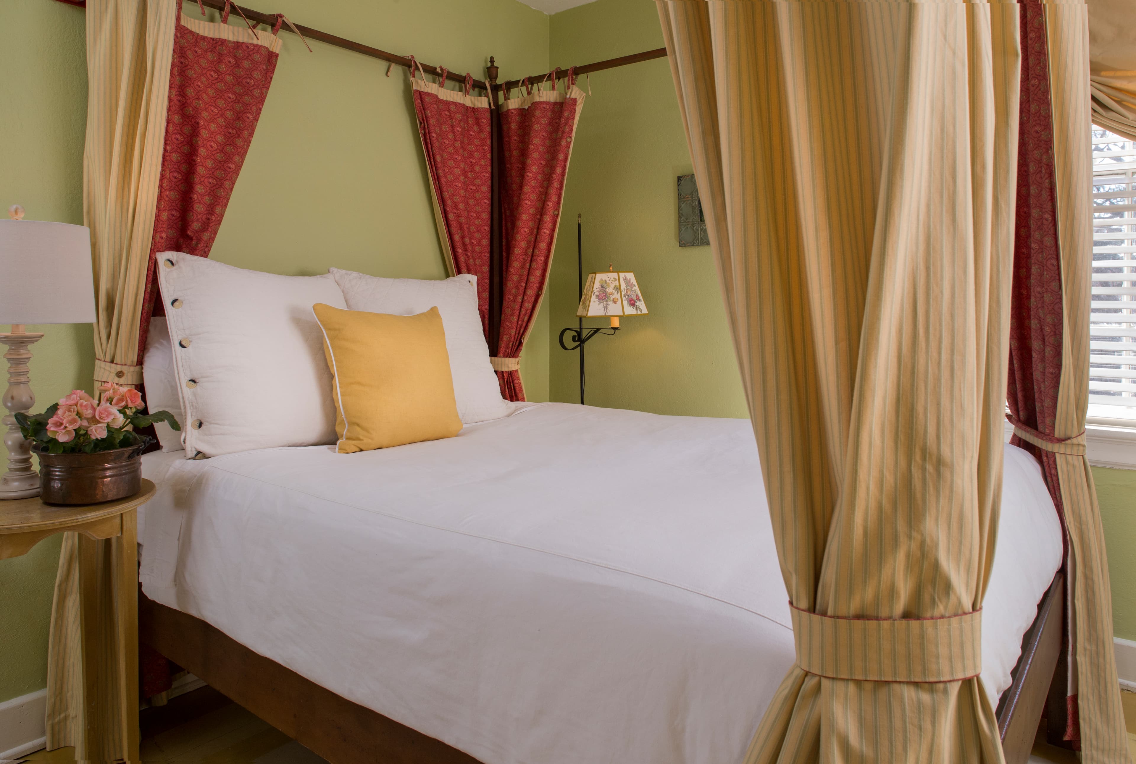 Four-poster canopy bed with red and gold drapes, nightstand with table lamp and flowers
