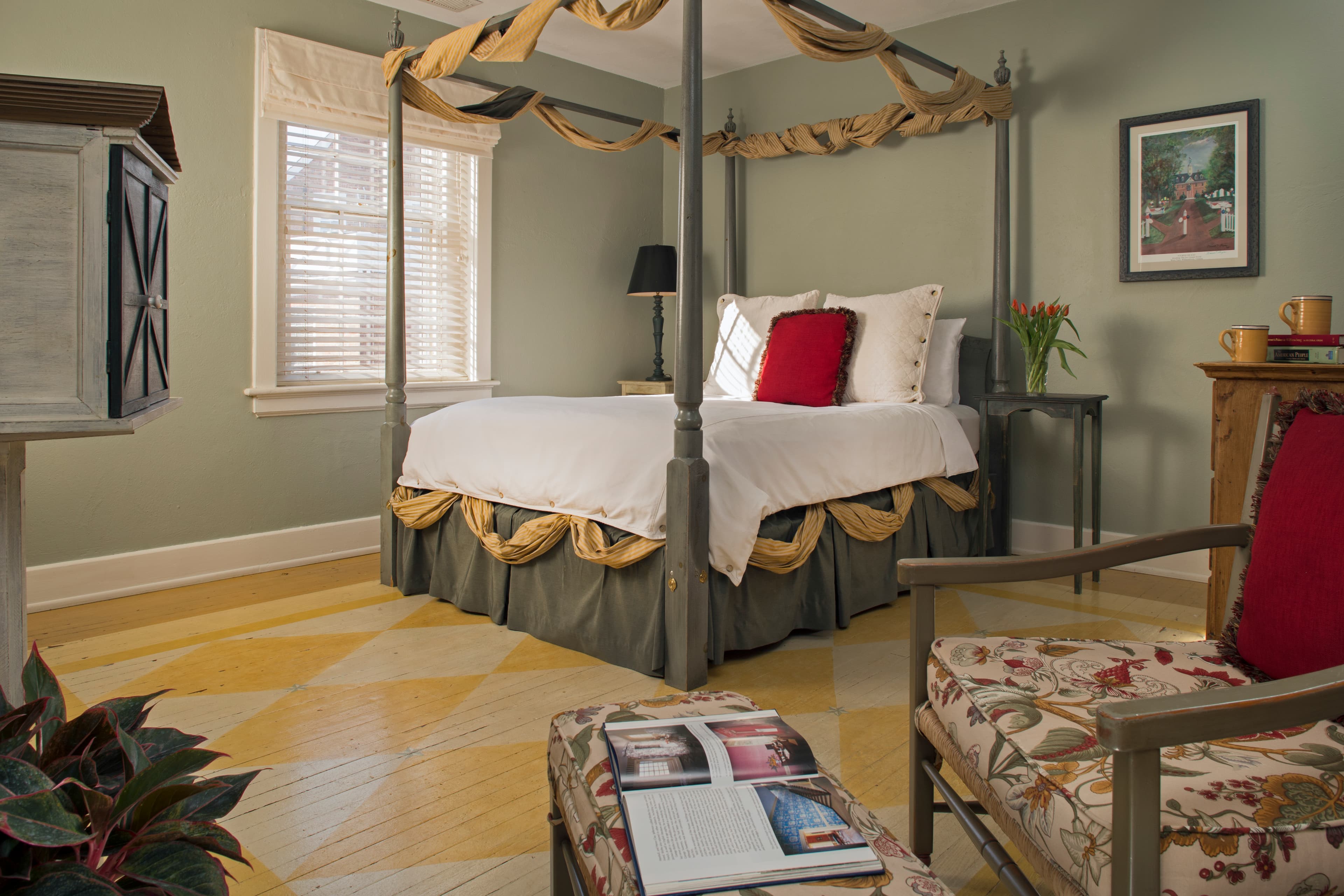 Painted wood four-poster bed swagged at the top with gold fabric, wooden chair with floral cushions and matching footstool in foreground