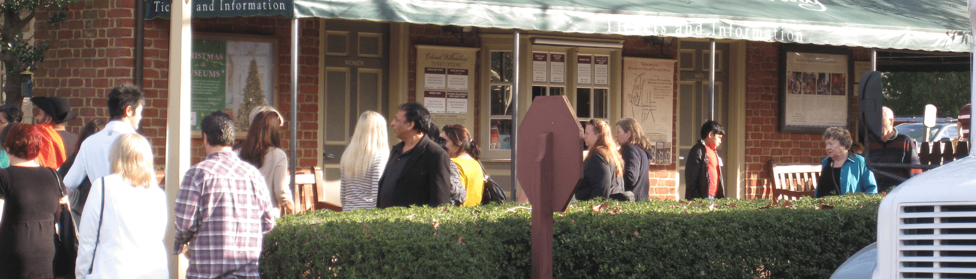 People standing around at a bus stop overhang with green shrubbery in the front.