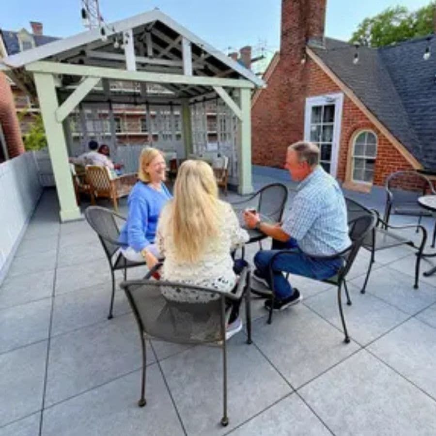 Three people converse on a rooftop terrace with a gazebo in the background.